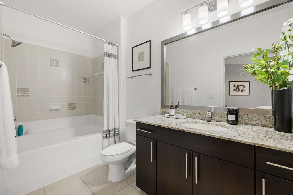 Modern bathroom with a bathtub-shower combo, white curtain, toilet, dark wood vanity with granite countertop, large mirror, and decorative art and flowers on the wall. The space is clean and well-lit.