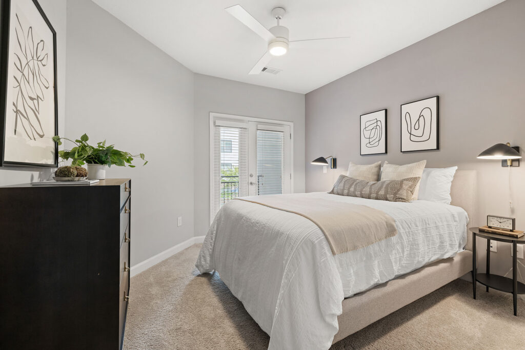 Modern bedroom with light gray walls, a large bed with white and beige bedding, minimalist art on the walls, a ceiling fan, dresser with a potted plant, and glass doors leading to a balcony.