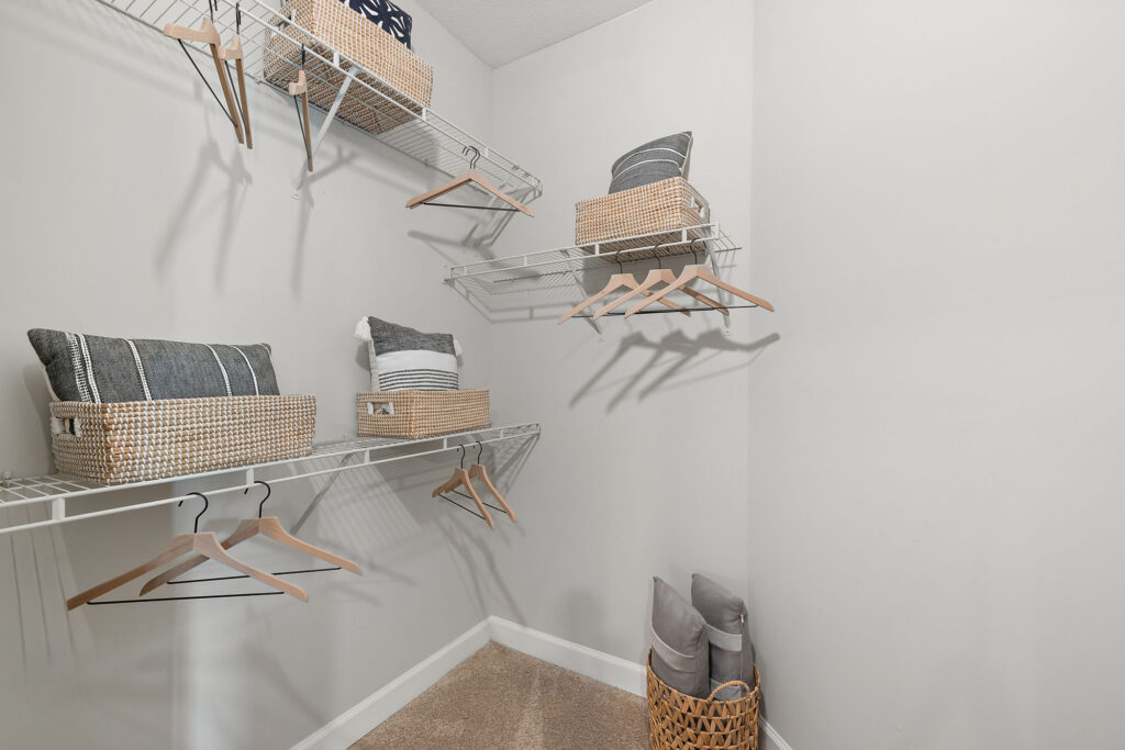 A walk-in closet with wire shelves holding baskets, folded pillows, and hangers. A woven basket with rolled towels sits on the carpeted floor. The walls are painted light gray.
