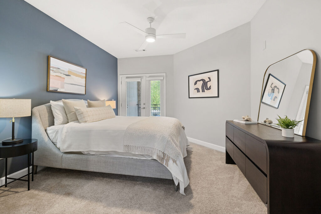 A modern bedroom with a blue accent wall, a large bed with white bedding, a nightstand with a lamp, abstract wall art, a dark dresser with a mirror, and glass doors leading to a balcony.