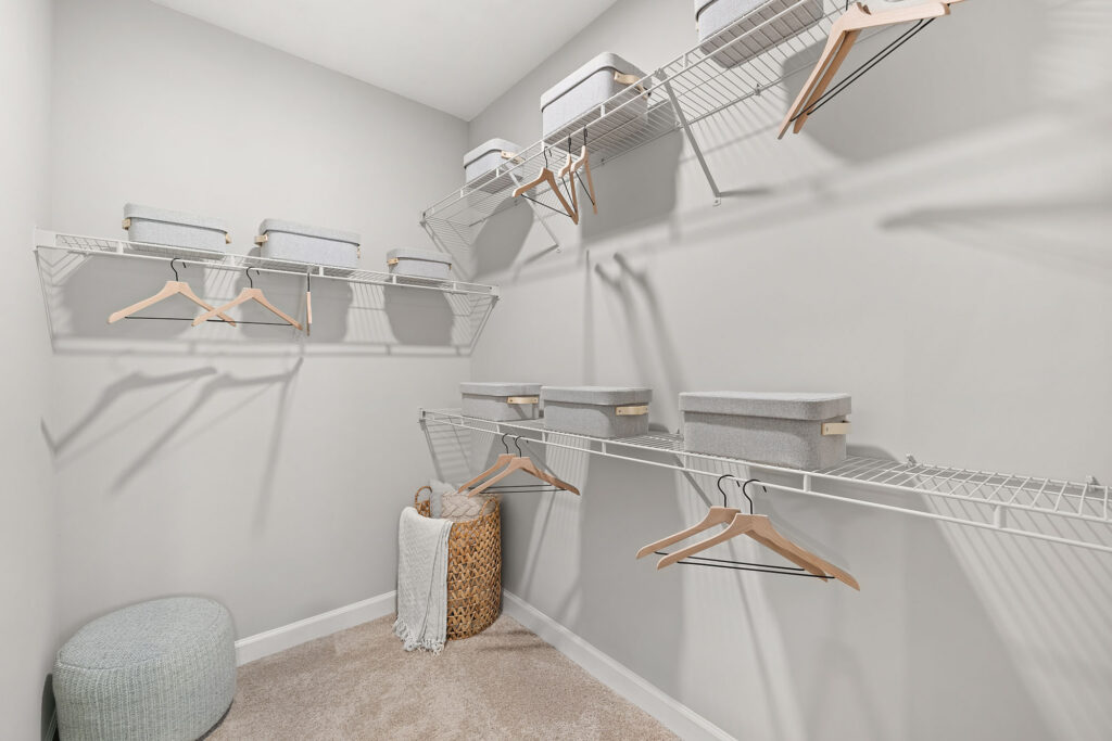 A spacious walk-in closet with white wire shelves holding gray storage boxes and wooden hangers, a woven basket with a blanket, and a light gray ottoman on a beige carpeted floor.