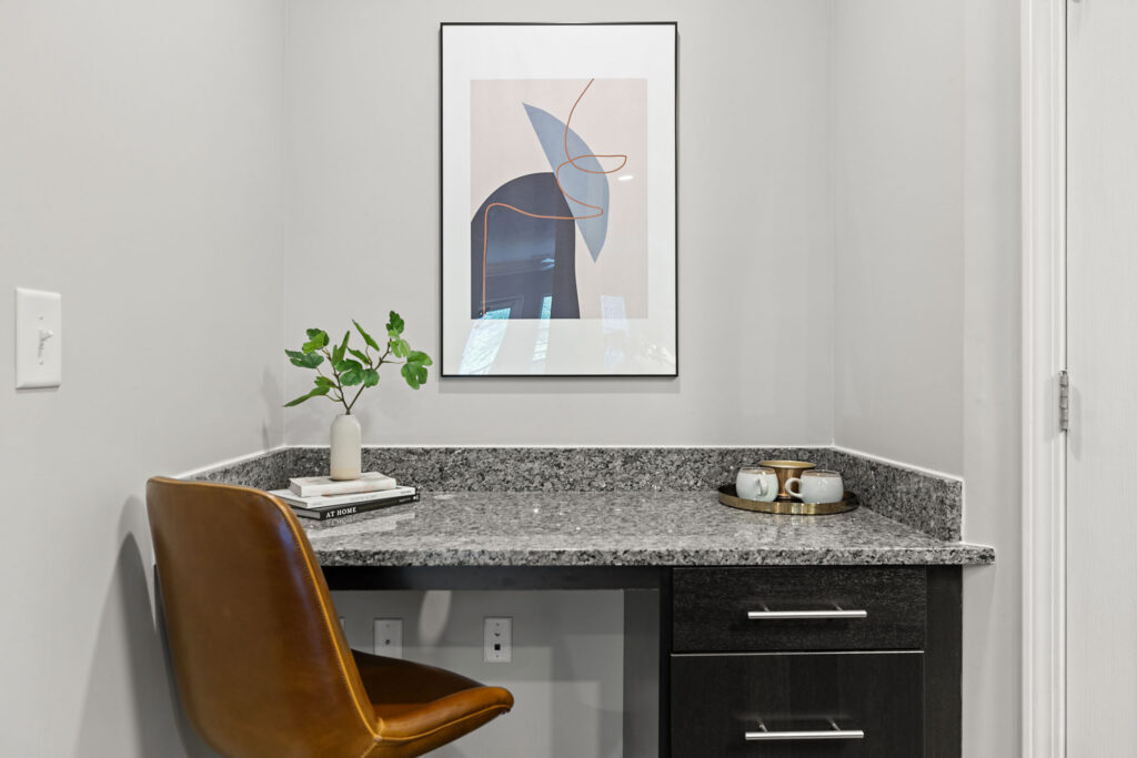 A small home office nook with a granite desk, brown leather chair, modern abstract art on the wall, a vase with greenery, a stack of books, and a tray with cups on the counter.
