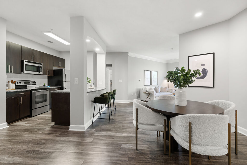 Modern open-concept apartment with a kitchen featuring dark cabinets and stainless steel appliances, a dining area with a round table and white chairs, bar seating, and a bright living area with minimalist decor.