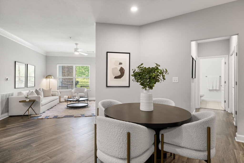 Modern open-concept living and dining area with light gray walls, wooden floors, a round dining table with four chairs, a cozy sofa, large window, wall art, and a potted plant centerpiece.