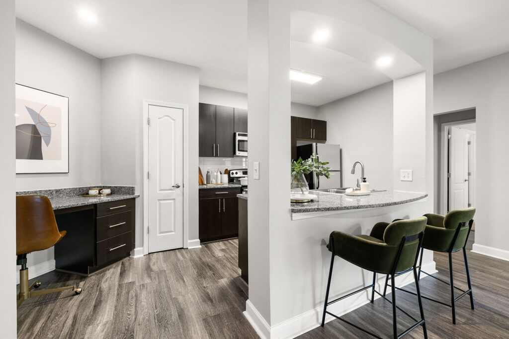 Modern kitchen with dark cabinets, stainless steel appliances, gray countertops, and a breakfast bar with two green velvet stools. An adjacent small office nook features a chair, desk, and abstract artwork.