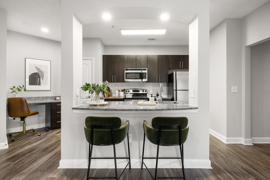 Modern kitchen with dark cabinets, stainless steel appliances, and a granite countertop breakfast bar with two green velvet stools. An adjacent workspace with a desk and chair is visible on the left. The space is well-lit and stylish.