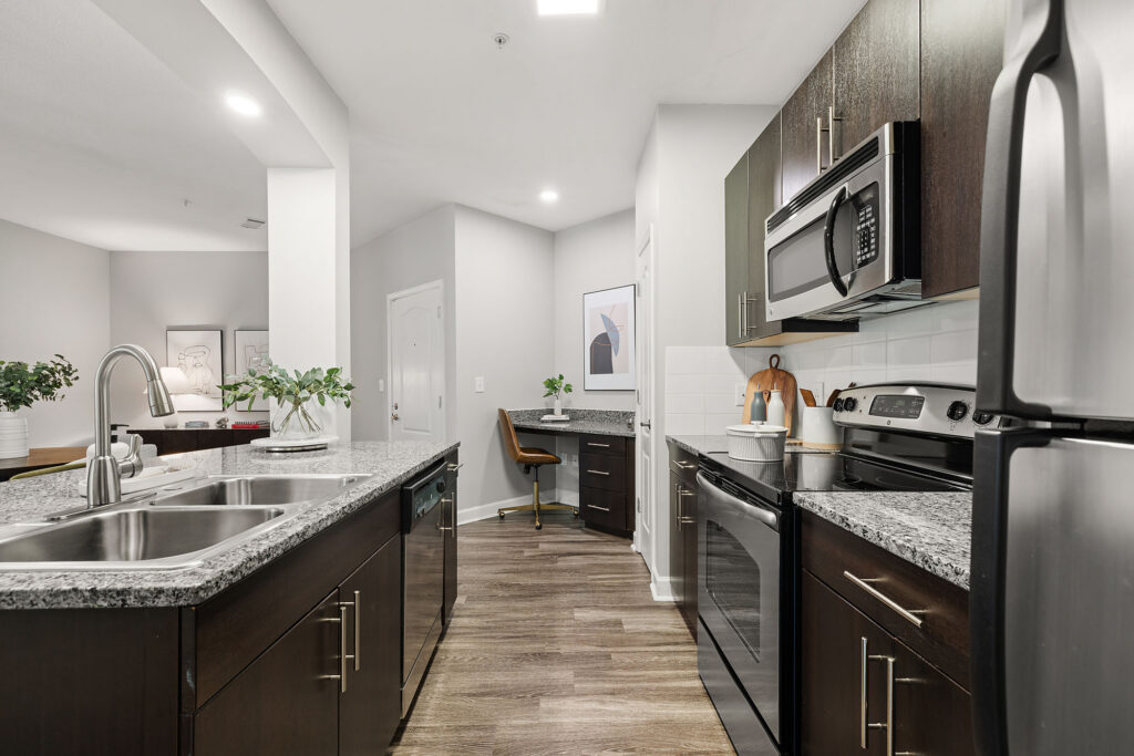 Modern kitchen with dark cabinets, stainless steel appliances, granite countertops, double sink, and hardwood floors; a small desk area with a chair and framed artwork is visible in the background.