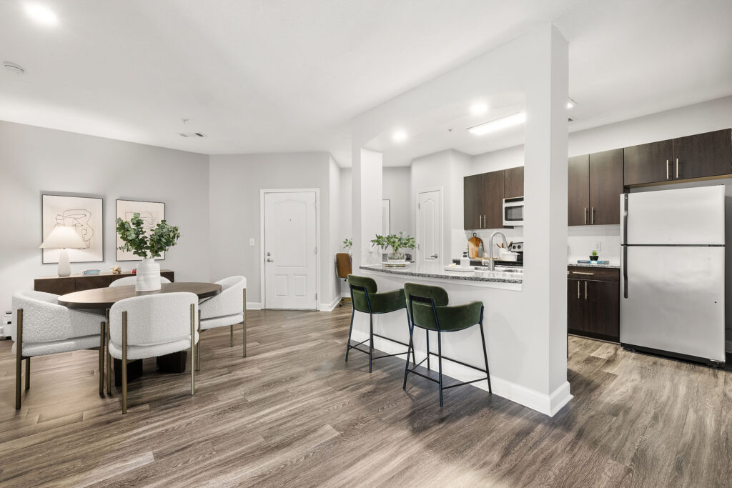 Modern open-concept apartment with a kitchen featuring dark cabinets and stainless steel appliances, a breakfast bar with green stools, a round dining table with white chairs, and light wood flooring throughout.