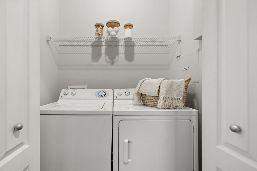 A compact laundry room with a white washing machine and dryer side by side. A basket with towels sits on the dryer, and jars are on a wire shelf above. The walls and doors are white.