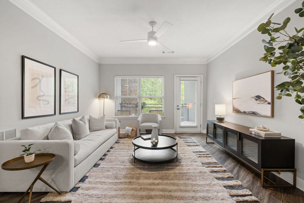 A modern living room with a light-colored sofa, abstract wall art, a round coffee table, a striped rug, a console table with decor, large windows, and indoor plants. The space is bright and neatly arranged.