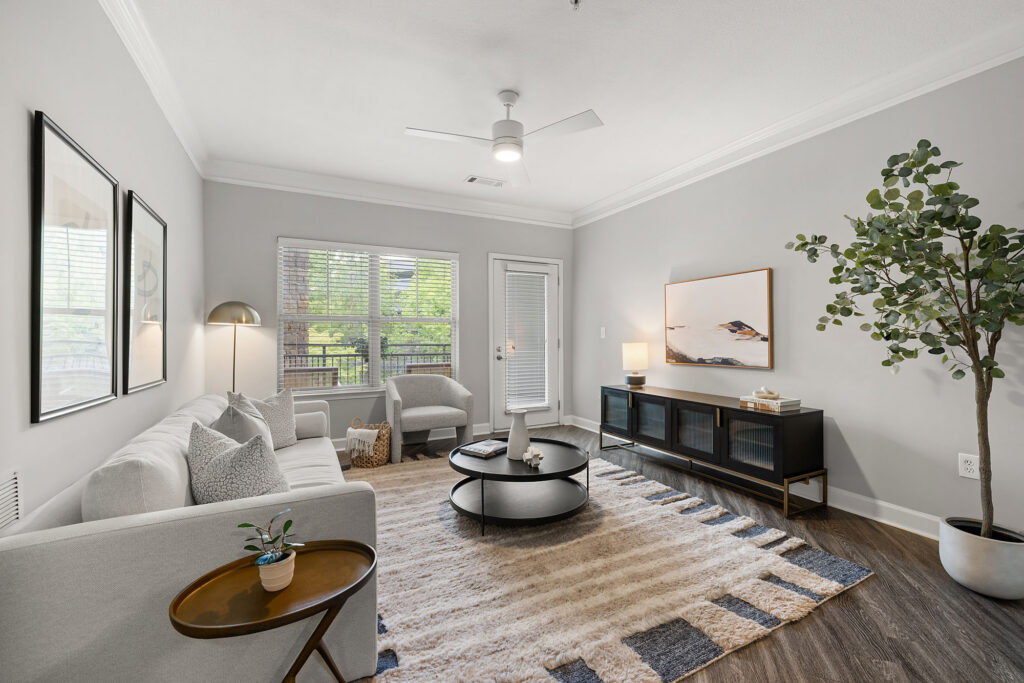 Modern living room with light gray walls, a white sofa, armchair, coffee tables, a TV console, large window, glass door, striped rug, indoor plant, and minimalist decor, all featuring neutral colors and natural light.