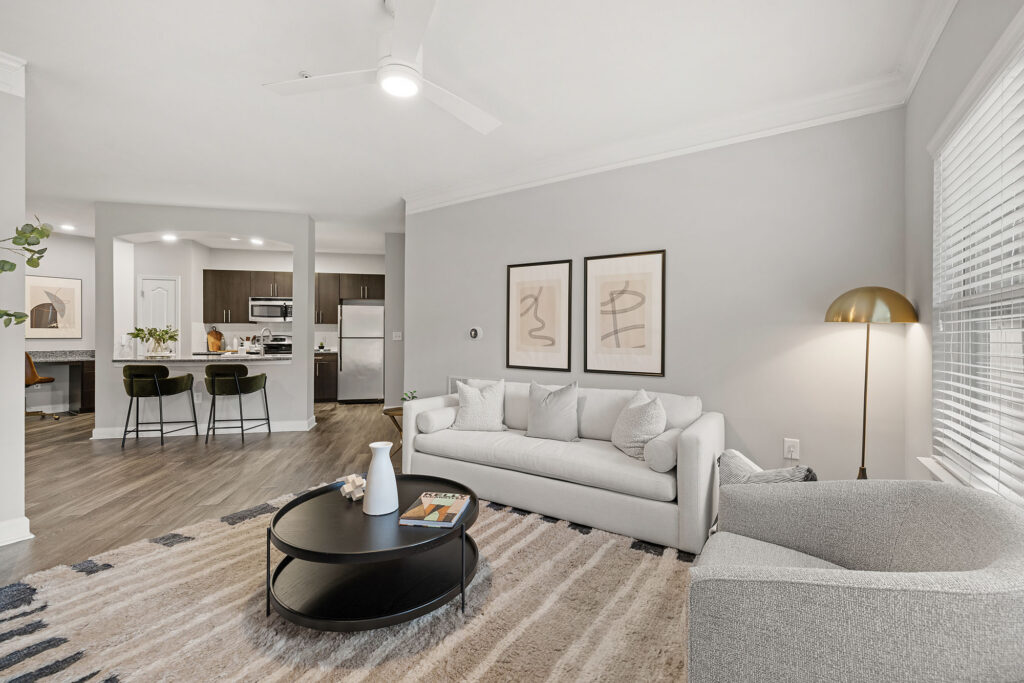 Modern living room with a light gray sofa, armchair, round black coffee table, abstract wall art, gold floor lamp, and open view of a kitchen with barstools and stainless steel appliances. Natural light from a large window.