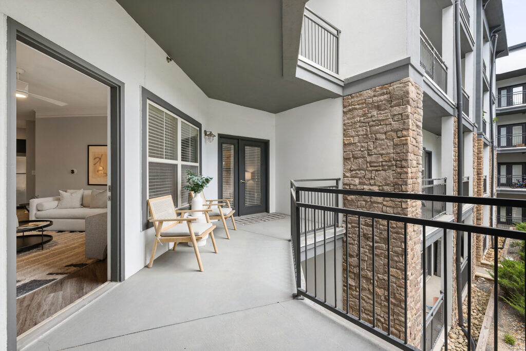 Spacious apartment balcony with two wooden chairs and a potted plant, adjacent to a living room with modern decor, large windows, and stone accent walls on the building exterior.