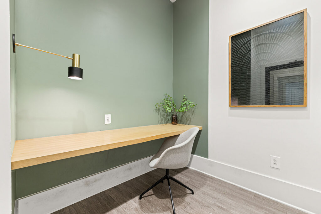 A minimalist workspace with a light wood floating desk, gray swivel chair, green accent wall, wall-mounted lamp, framed abstract art, and a small leafy plant in a vase on the desk.