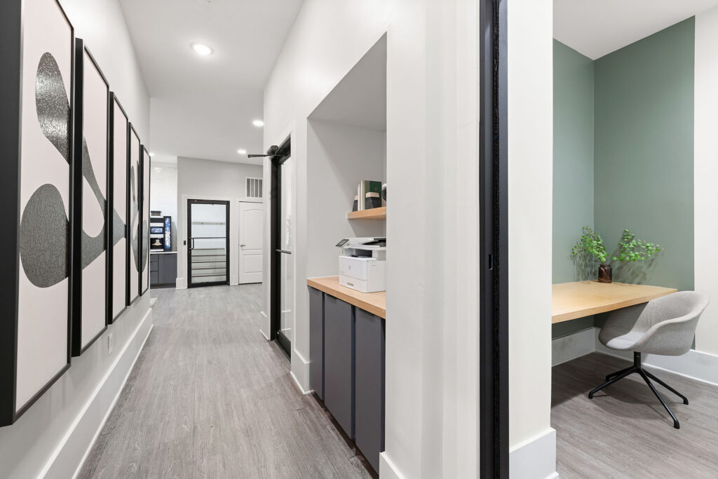 A modern office hallway with abstract art on the wall, light wood flooring, a built-in printer station, and a small workspace with a desk, chair, and green accent wall with a potted plant.