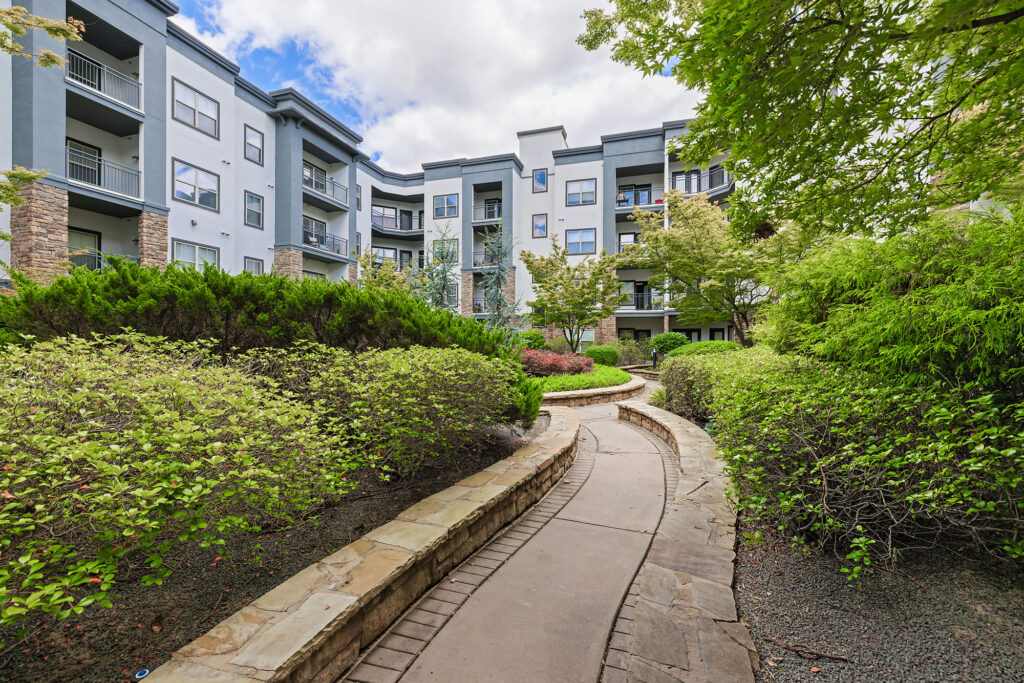A landscaped courtyard with lush green bushes and trees, featuring a winding stone-lined pathway leading to a modern multi-story apartment building with balconies under a partly cloudy sky.