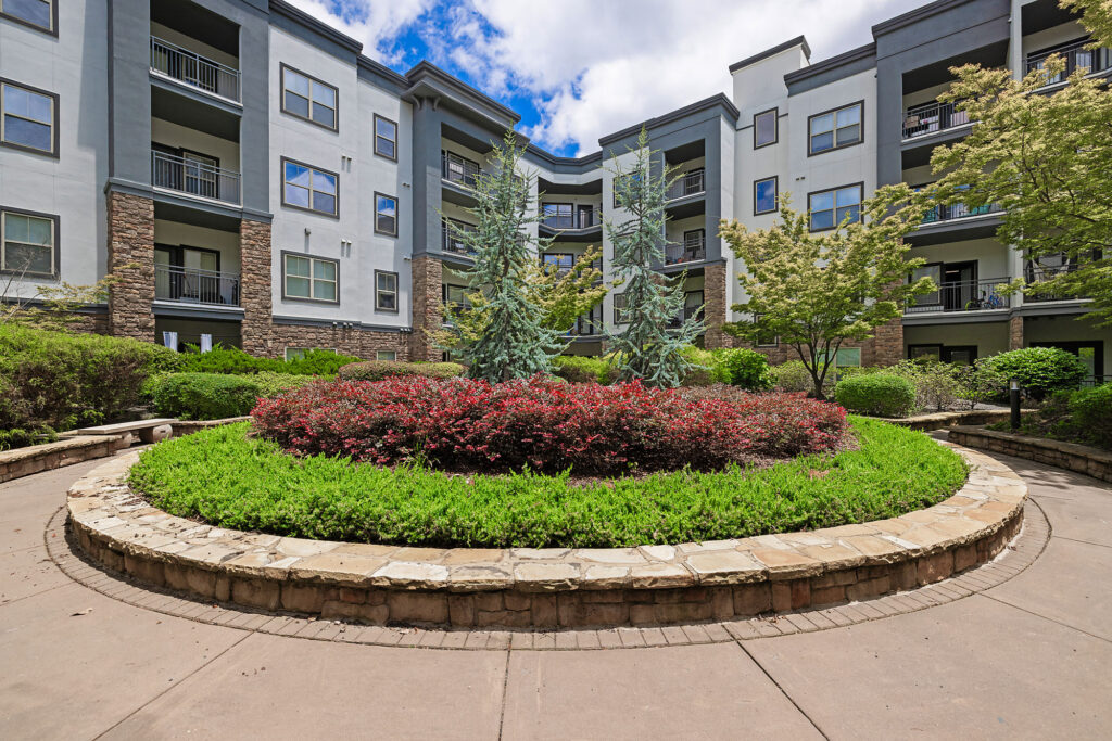 A landscaped courtyard with a circular stone planter featuring shrubs, small trees, and greenery, surrounded by a modern multi-story apartment building under a partly cloudy sky.