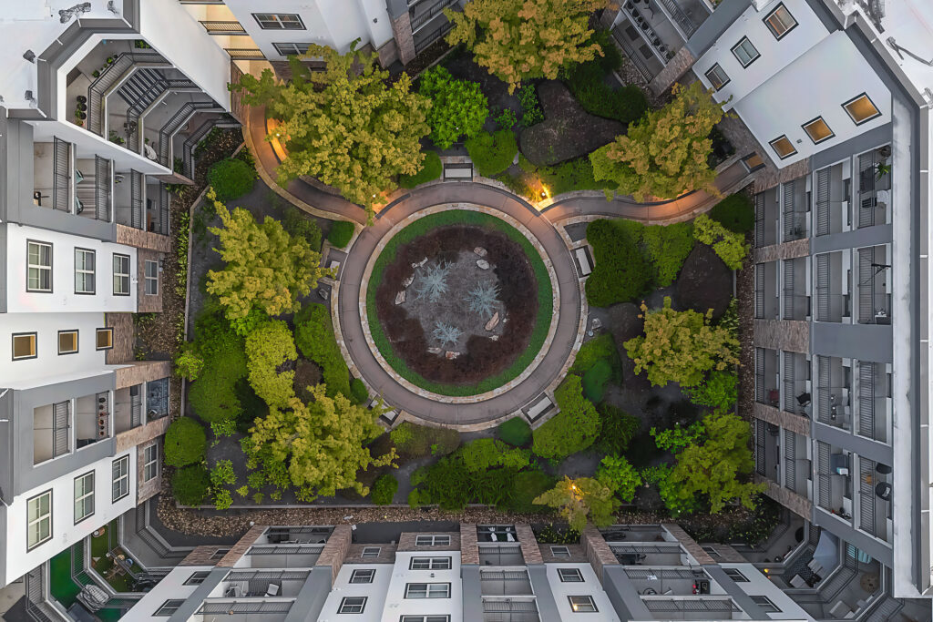 Aerial view of a modern apartment building courtyard with a circular garden in the center, surrounded by trees, benches, pathways, and illuminated by warm lights.